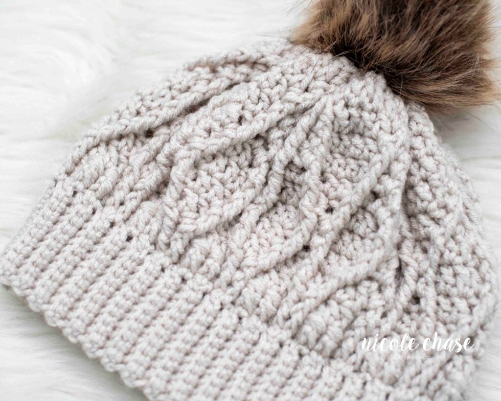 Close up of the Adaleigh Beanie in a cream color, detailing the textured cables and a brown faux fur pom.