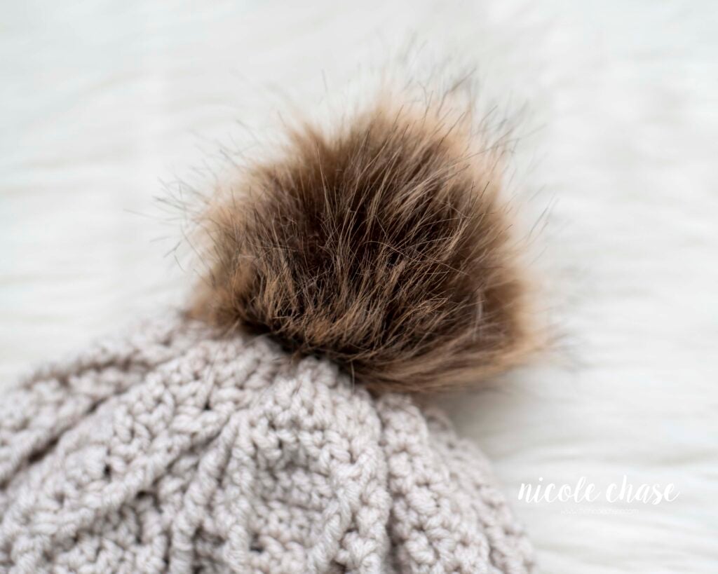 A cream-colored textured crochet beanie with a close up on the brown faux fur pom pom. This free crochet toque pattern is shown in a flat-lay on a white faux fur background. 