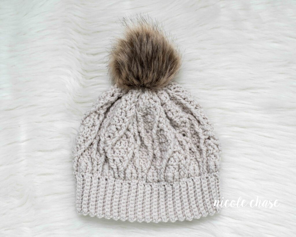 A cream Adaleigh Beanie with a brown faux fur pom, resting on a white faux fur surface. 