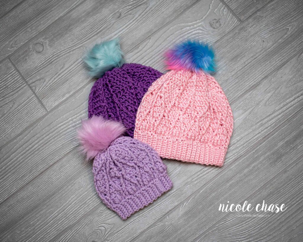 A flat lay of the Adaleigh Beanie made in lavender with a periwinkle pom, a deep purple with a light blue pom, and  a light pink with a multicolor pom, all made with the free beanie crochet pattern. 
