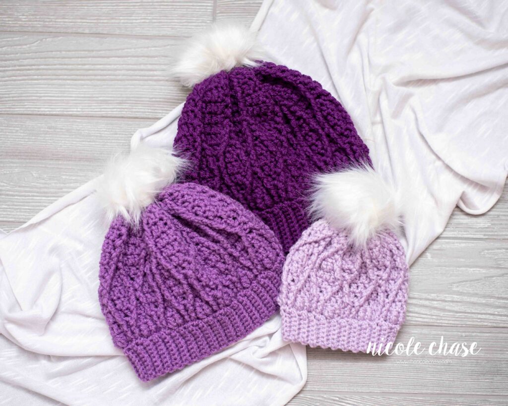Three Adaleigh Beanie's in various sizes, the smallest in a lavender color, the next in a purple color, and the largest in a deep purple color, all with white faux fur poms. 