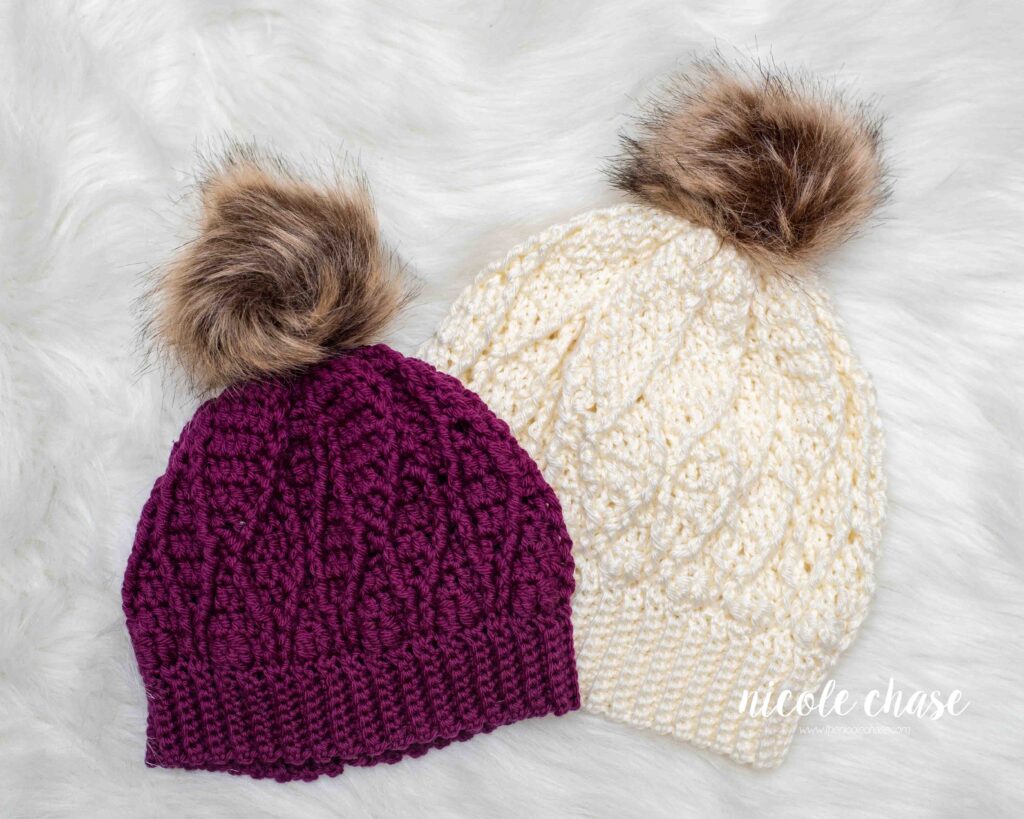 Two Adaleigh Beanie's, one in a deep burgundy color, and the other in a light cream color, both with a brown faux fur pom. 