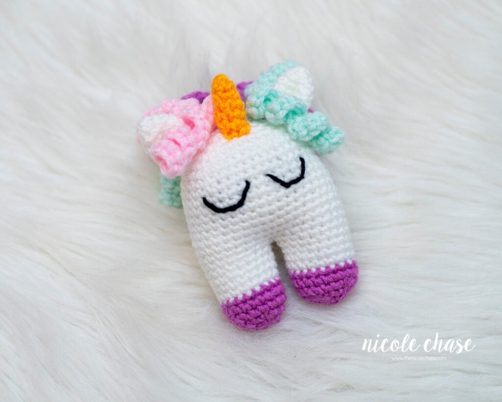 Crocheted Elanna the Unicorn baby toy resting on a white fluffy surface, displaying embroidered sleeping eyes, colorful pastel curls, and purple hooves.