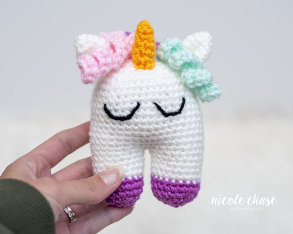 Front view of small crocheted Elanna the Unicorn baby toy held by the leg, showing its embroidered sleeping eyes, orange horn, and pastel pink, mint, and purple accents.