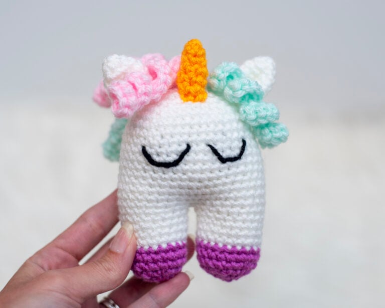 A close-up of the small, crocheted Elanna the Unicorn baby toy, featuring a white body, embroidered sleeping eyes, colorful pastel curly hair, and purple hooves, held in a hand.