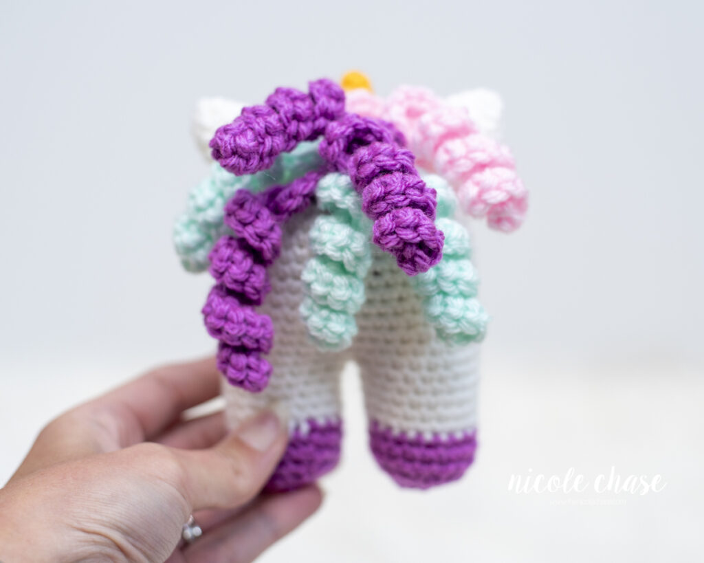 Back view of the crocheted Elanna the Unicorn baby toy held in a hand showing the colorful purple, mint, and pink spiral curls attached for the mane.