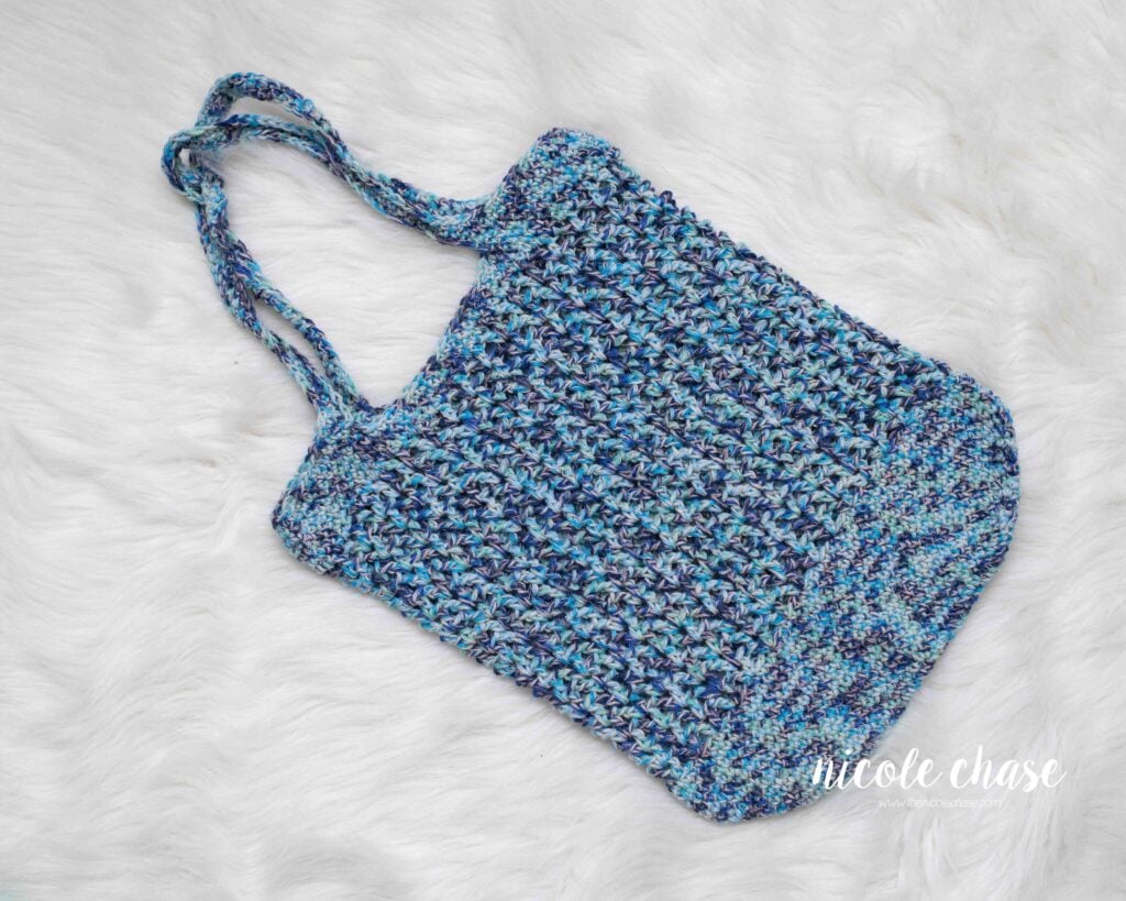 Blue multi-tonal crochet market bag with an airy V stitch mesh design, laying on a white fur background. 
