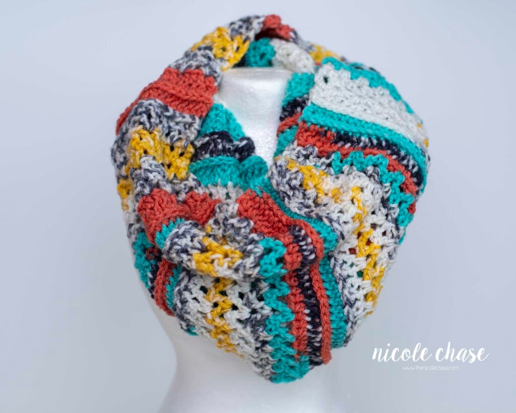 A multi-colored crochet cowl (New Beginnings Cowl) in orange, teal, yellow, and white, displayed on a mannequin. Get the free cowl crochet pattern here!