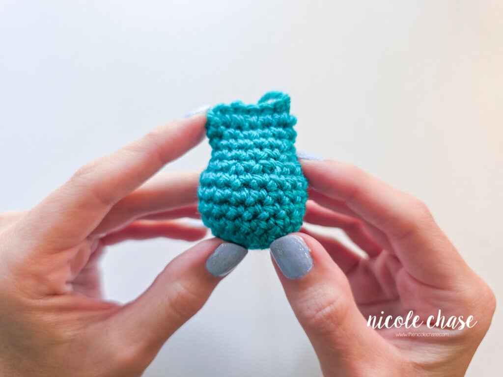 A hand holding a small, teal crocheted amigurumi demonstrating a smooth finish made with an invisible decrease.