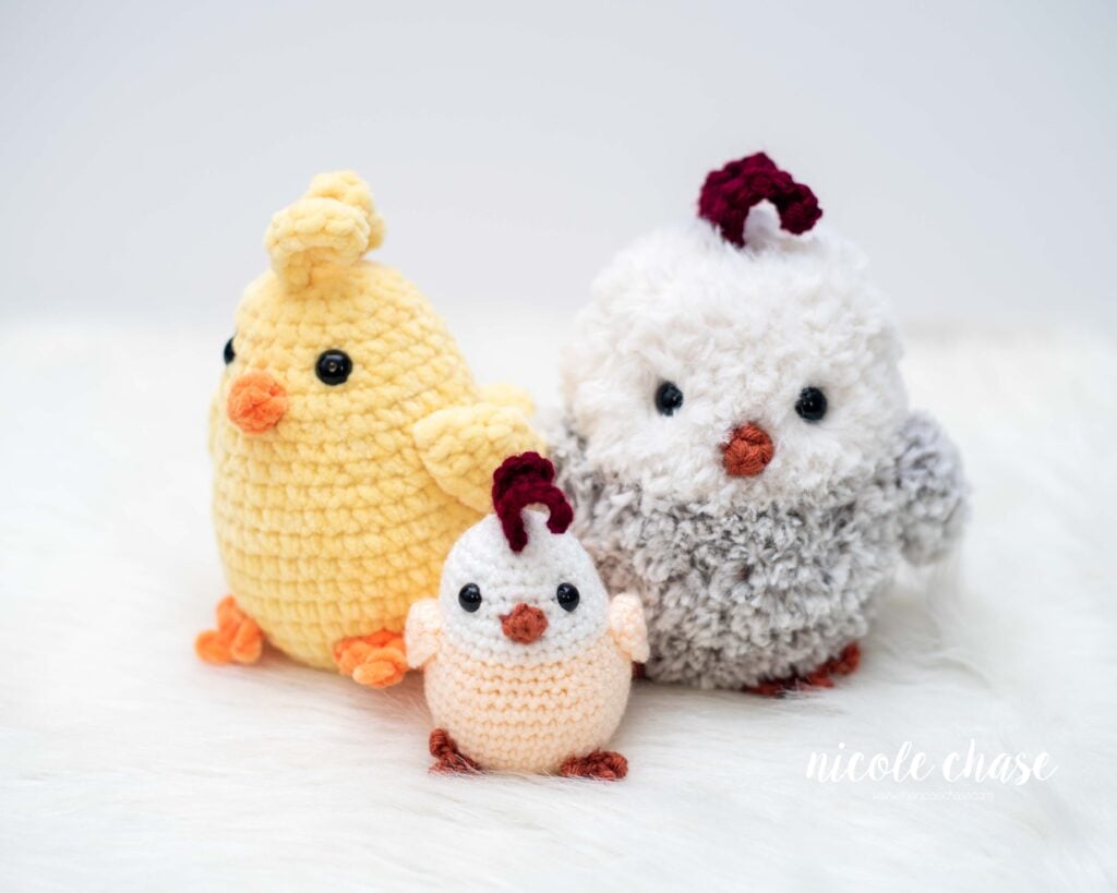 Group photo of three adorable crochet chicken amigurumi toys sitting on a white background. The smallest chicken is cream and white, the medium sized chicken is bright yellow to look like a chick, and the largest chicken is fluffy white and gray, demonstrating how different yarn weights can be used for the free chicken crochet pattern. 