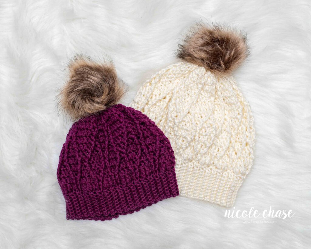 two textured cable hats with matching brown faux fur poms, shown in a magenta color and a neutral cream color