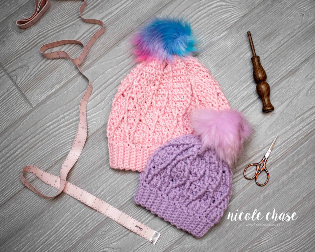 pink, child size, crochet cable hat with rainbow faux fur pom with a purple, infant size, crochet cable hat with purple faux fur pom, amongst craft supplies
