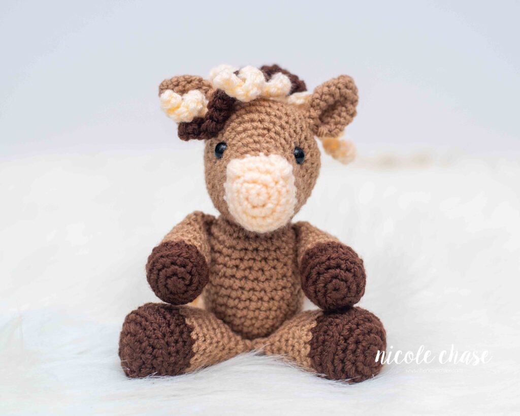 small, light brown, crochet horse amigurumi with dark brown hooves and a cream accent color, in a seated position
