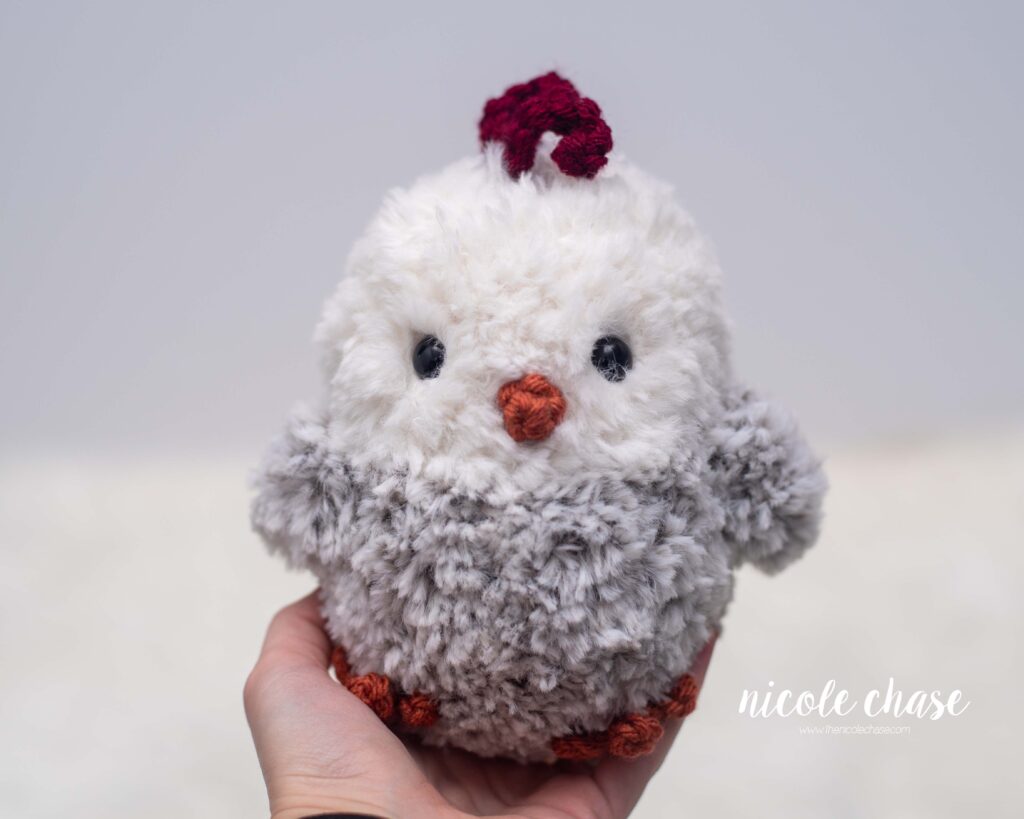 crochet chicken made with grey and cream fur yarn, sitting on a hand