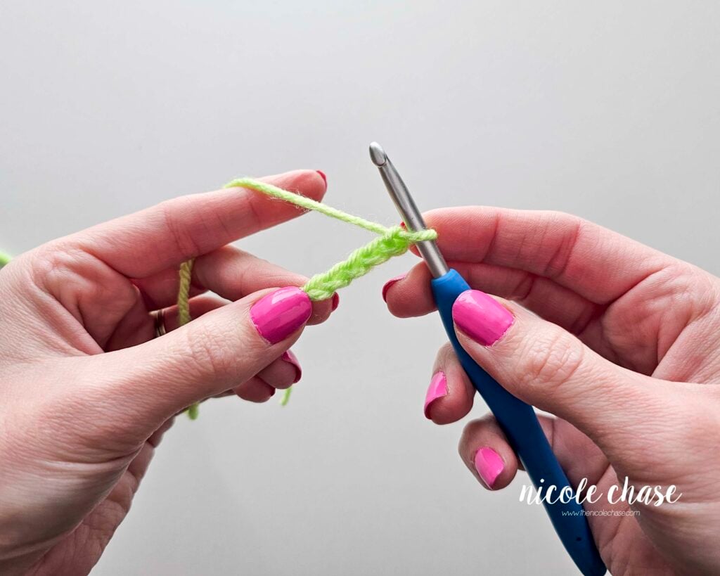 Close up of a beginner crocheter's hands holding a crochet hook and working a crochet chain stitch using bright green yarn for the tutorial on creating a slip knot and how to crochet a chain stitch.