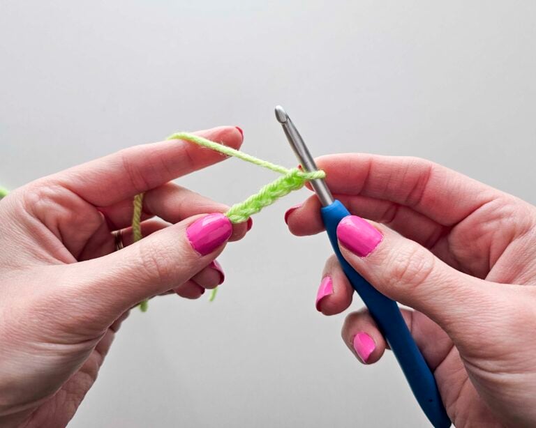 Hands holding a blue crochet hook and working a chain stitch in a bright green yarn, demonstrating this slip knot and chain tutorial.