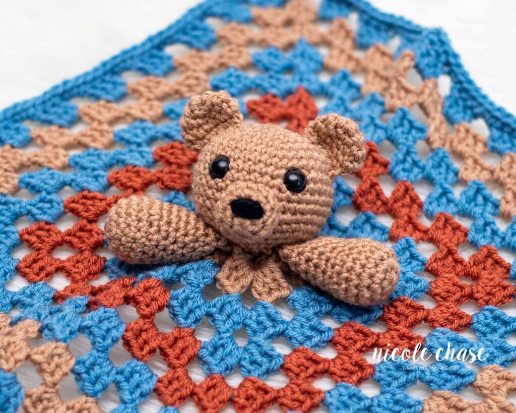 Close-up of the finished teddy bear lovey crochet pattern head and arms, resting on the blue, brown, and rust granny square crochet lovey blanket. This is a free bear security blanket pattern.