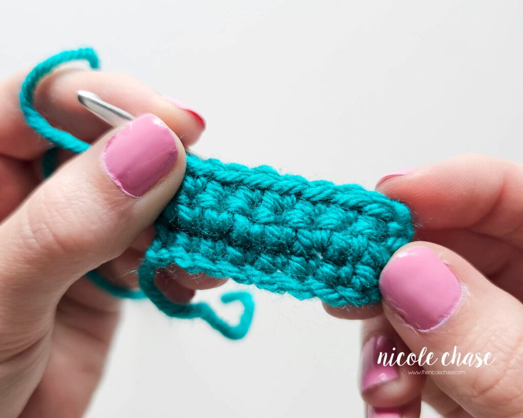 Front view of a teal single crochet swatch showing the uniform single crochet stitches, held with a crochet hook to demonstrate the single crochet (SC) technique for beginners.