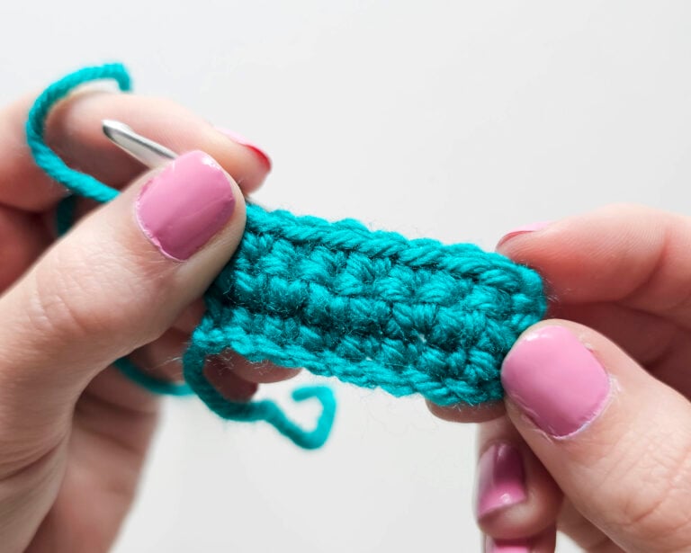 A close-up view of a hand holding a teal crochet swatch, demonstrating the texture of several rows of single crochet stitches with a silver crochet hook.