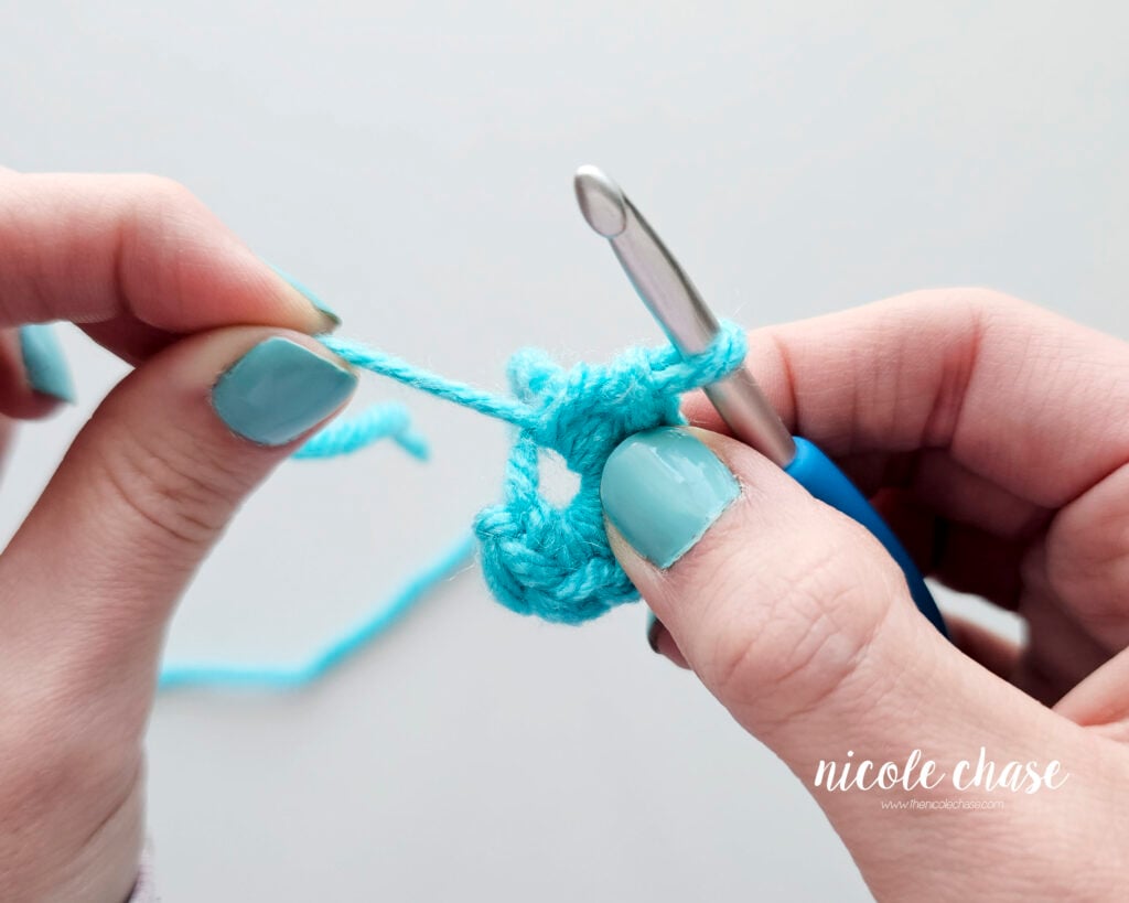 A close-up photo of a person's hands pulling the yarn tail of a crochet magic ring to close the center hole. The image shows blue yarn and a blue crochet hook, demonstrating how to adjust a magic circle for amigurumi, part of the how to make a magic ring tutorial.