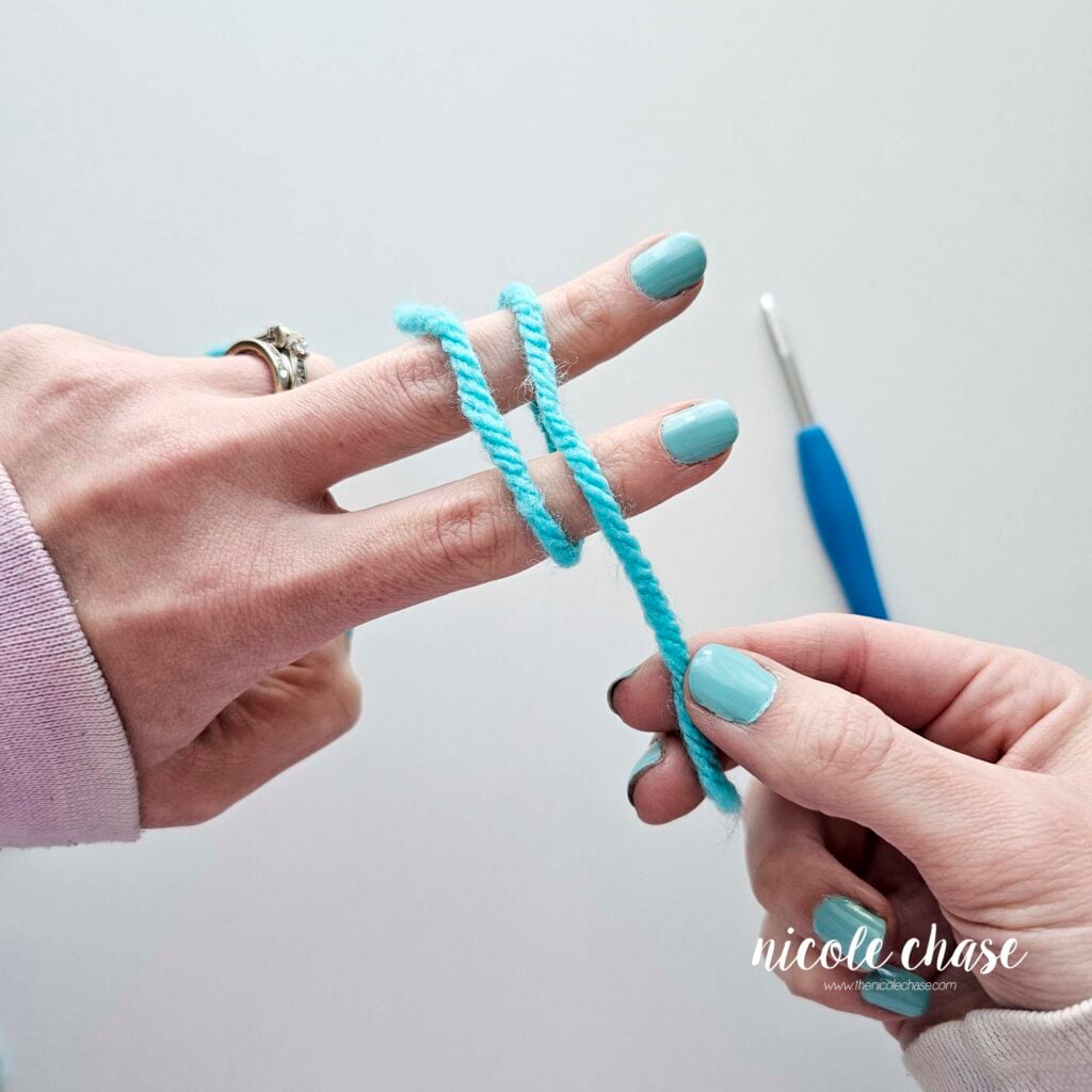 Close-up showing the person wrapping the blue yarn around the extended fingers. The crochet hook is visible in the background.