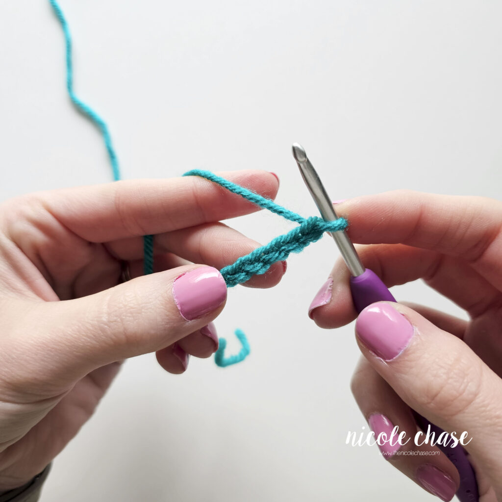 Close-up of hands holding teal yarn in a chain of stitches on a crochet hook.