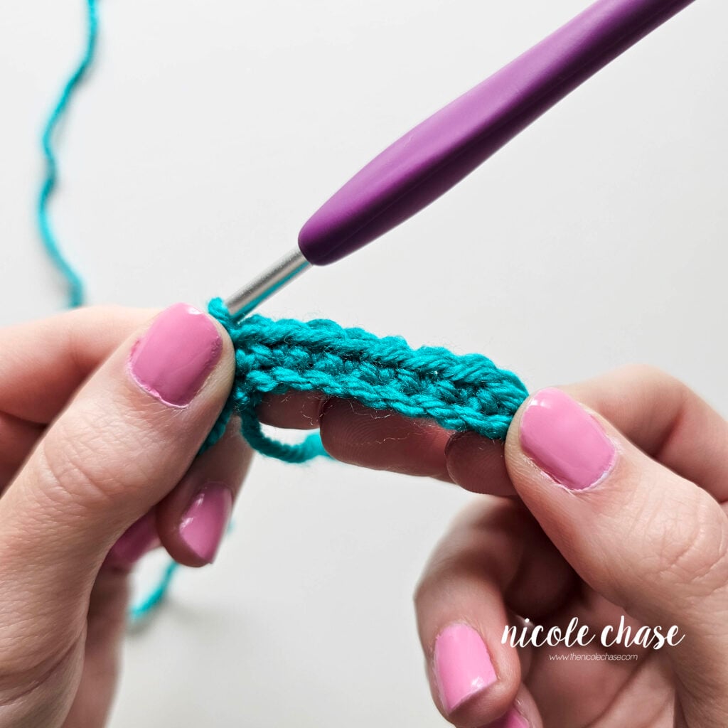 A completed row of single crochet stitches across a small chain made in teal yarn and being held in hands with a crochet hook.