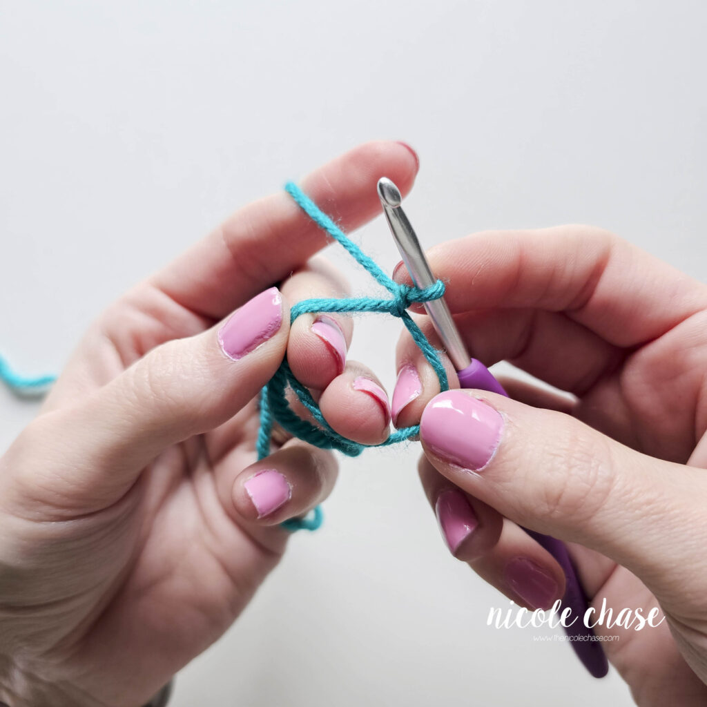 A close-up of hands holding teal yarn in a magic ring, attached to a crochet hook. The magic ring is open and ready for stitches.