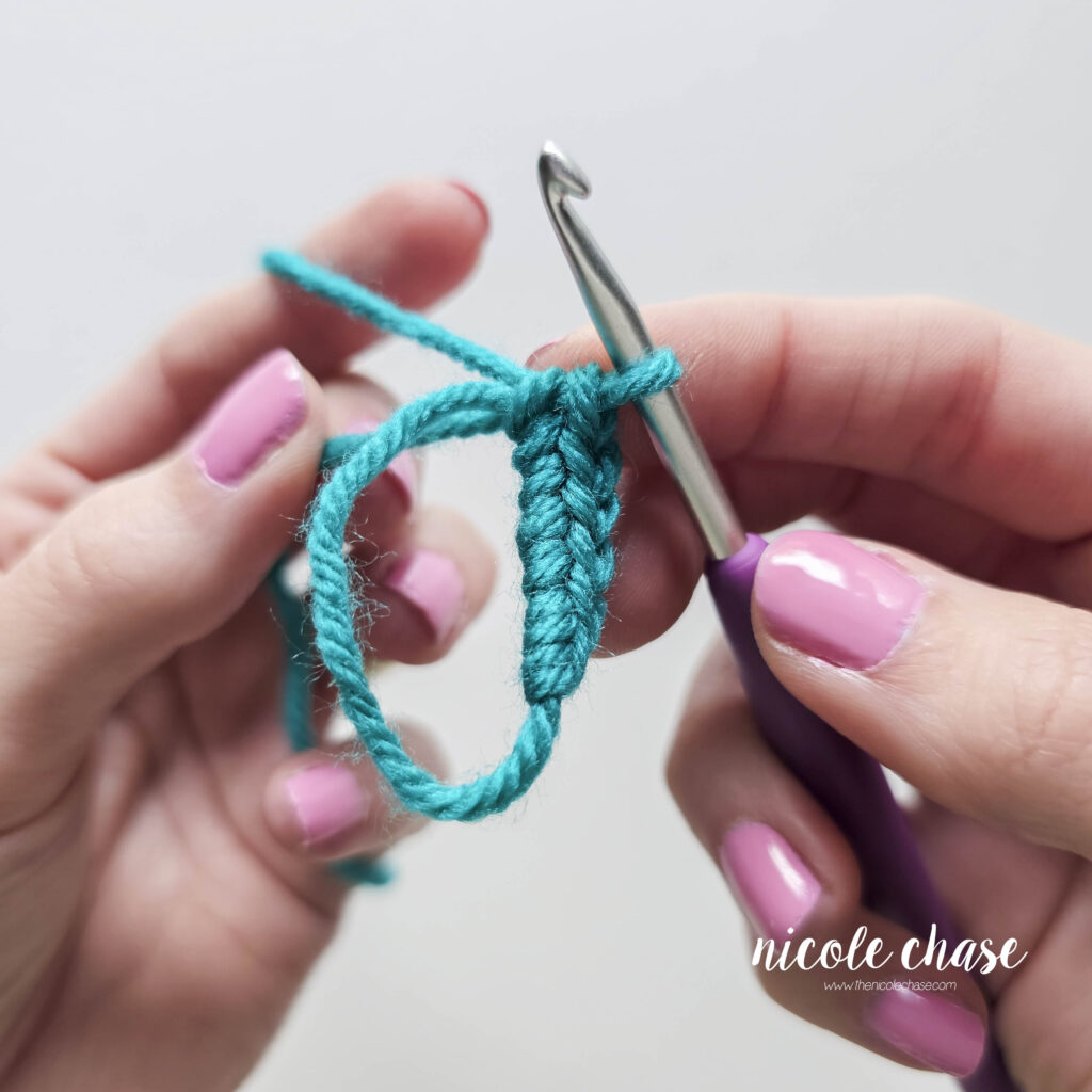 A close up of a crochet magic ring with 6 single crochet stitches worked into the ring. One hand is holding the hook while the second hand is pulling the yarn tail to tighten the magic ring.