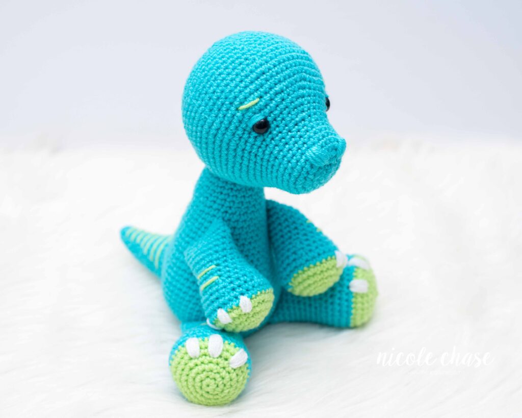 A sitting teal crocheted apatosaurus dinosaur with lime green accents on the feet and white embroidered nails. 