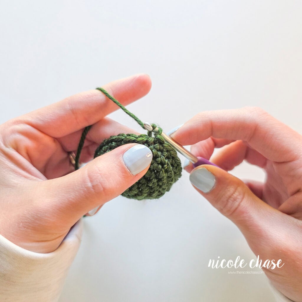 Yarning over with the cut yarn tail using a purple crochet hook on a green crocheted piece to fasten off crochet. 