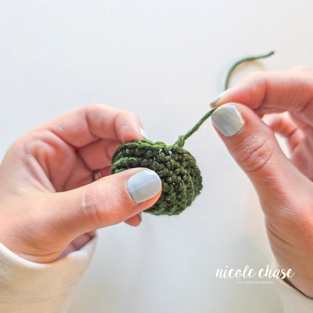 Hands pulling on the yarn tail to tighten the fasten off knot on a green amigurumi piece. 