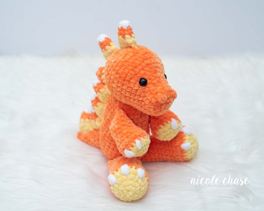 Side angle view of Mini Toby the Tarbosaurus plush sitting on white fur, featuring orange chenille yarn, yellow feet, and candy corn spikes.