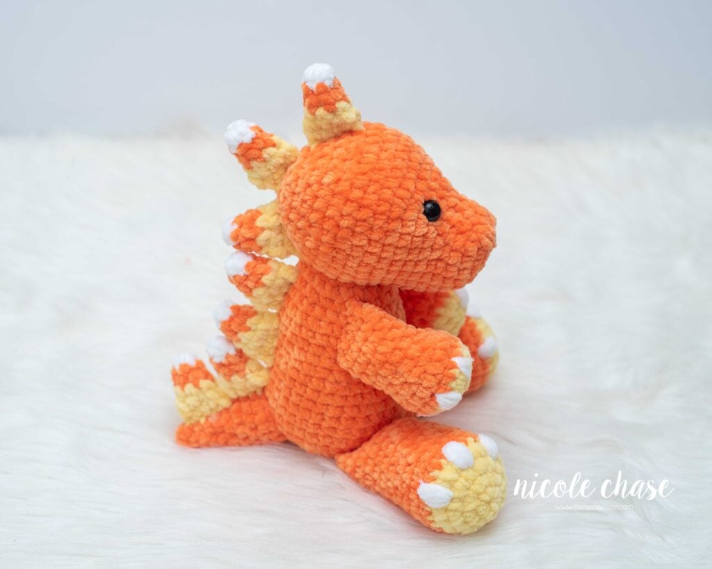Side profile view of Mini Toby the Tarbosaurus crocheted in orange plush yarn, showing a row of candy corn colored spikes along the back and tail.