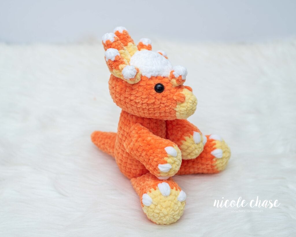 Side angle view of Mini Stevie the Stygimoloch plush sitting on white fur, highlighting the white head dome, yellow snout, and candy corn spikes.