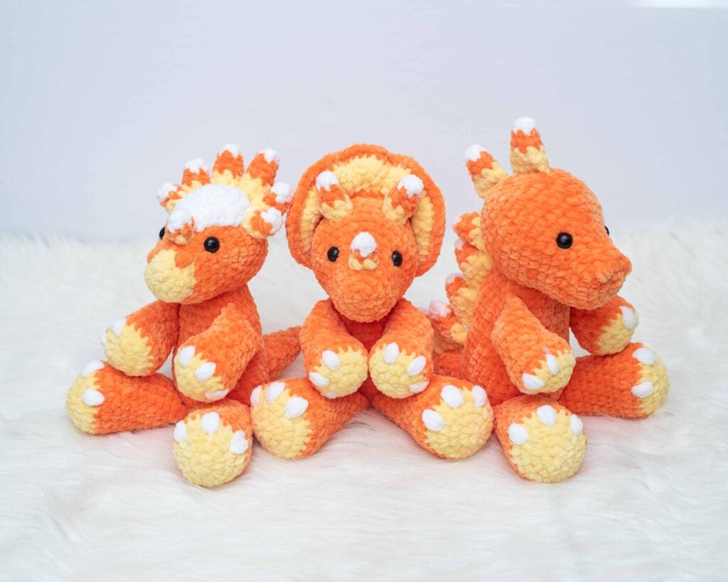 Three plush crocheted candy corn dinosaurs - a Stygimoloch, Triceratops, and Tarbosaurus, made with orange, yellow, and white plush yarn with candy corn details, sitting on a white surface.