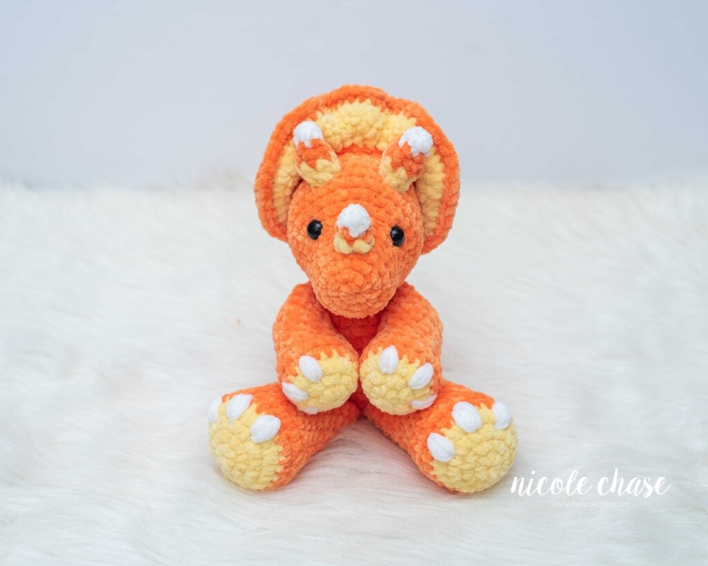 Front view of Mini Tanner the Triceratops crocheted in orange plush yarn with yellow feet and frill, featuring white and yellow striped candy corn horns.