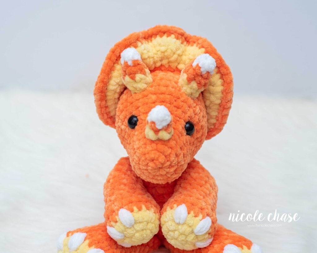 Close-up front view of Mini Tanner the Triceratops showing white, orange, and yellow candy corn horns, a yellow frill, and white nails on orange plush yarn.