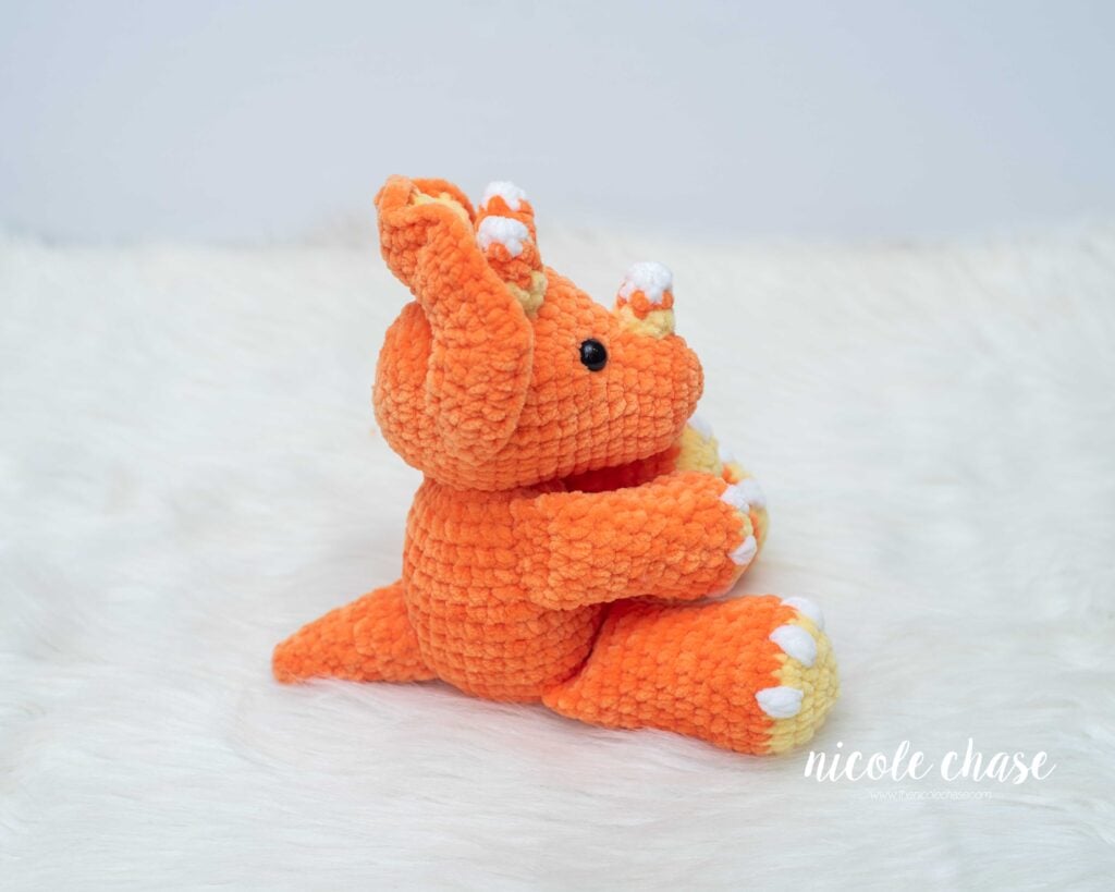 Side profile view of Mini Tanner the Triceratops made with orange plush yarn, showing the tail, frill profile, and white-tipped candy corn horns.