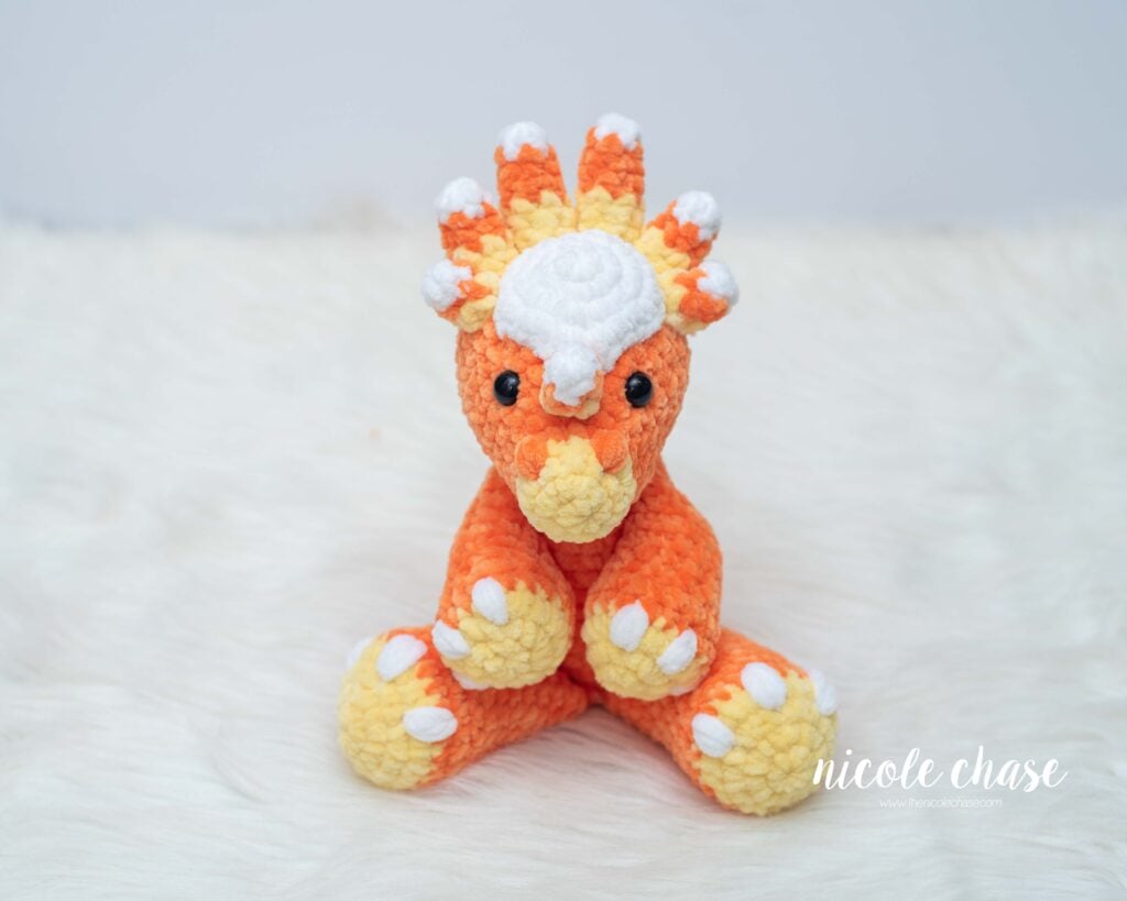 Front view of Mini Stevie the Stygimoloch crocheted in orange plush yarn, featuring a white head dome and candy corn striped spikes around the head.