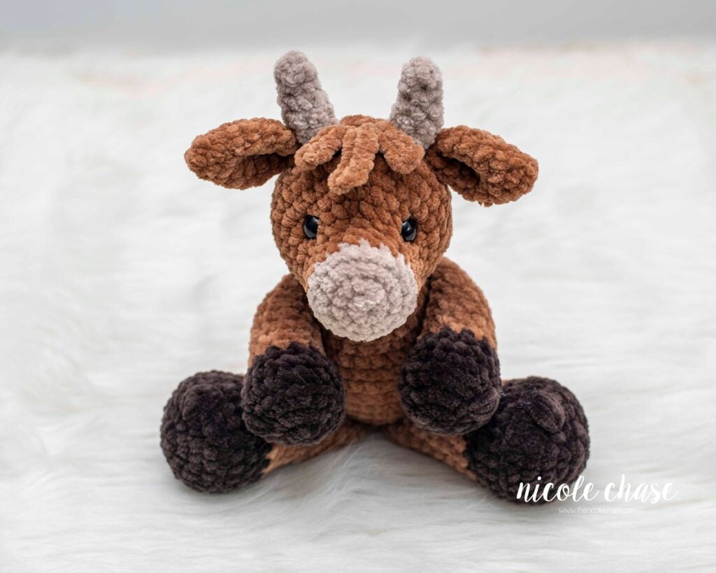 A handmade plush crochet highland cow sitting upright, featuring a medium brown body, dark brown hooves, a light tan snout and horns, a textured fringe hair, and black safety eyes, displayed on a soft white fur backdrop.