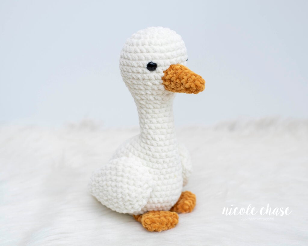 A handmade plush crochet goose sitting upright, featuring an off-white textured body, a gold beak, webbed feet, and black safety eyes, displayed on a soft white faux fur surface.