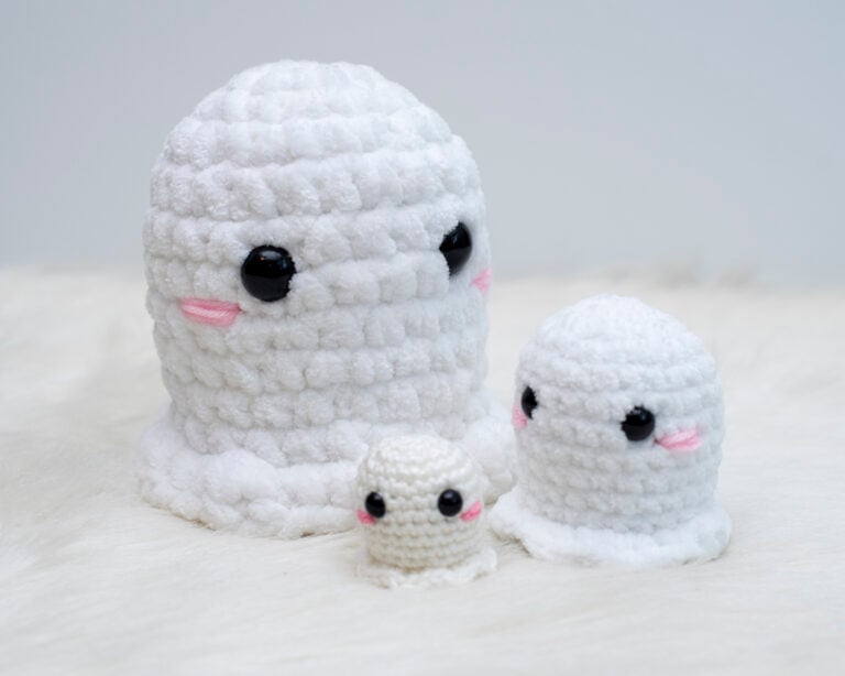 Three white crochet ghost amigurumi in different sizes made with worsted weight yarn and plush yarn sitting on a white furry background for a free crochet ghost pattern.