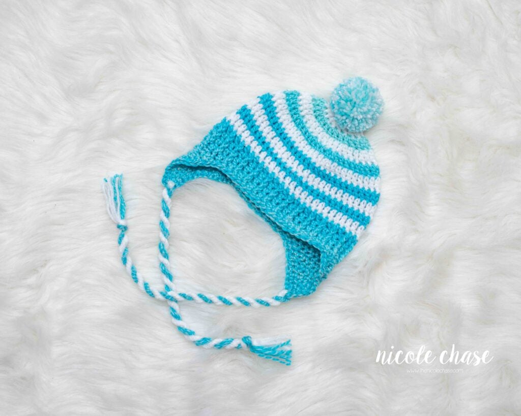 Finished earflap beanie crochet pattern shown in white and teal blue, with braided tassels and a pom pom on top.