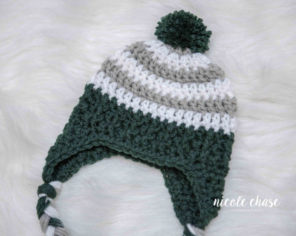 Close up of a baby sized Spirit Earflap Hat made in light gray and white stripes with a dark green brim and pom pom.
