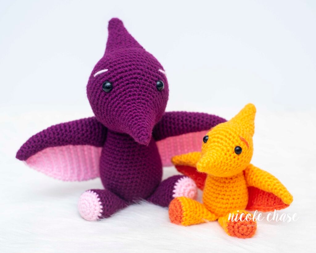 Side-by-side comparison of the full-sized purple Tori the Pterosaur sitting next to the small orange Mini Tori, showing scale for both free pterodactyl crochet patterns.