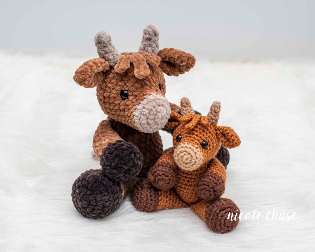Two amigurumi highland cows sitting on a white fur background. The left is larger and made with plush yarn, and the right is smaller and made with worsted weight yarn. Both are made with the Hazel the Highland Cow crochet pattern by Nicole Chase.