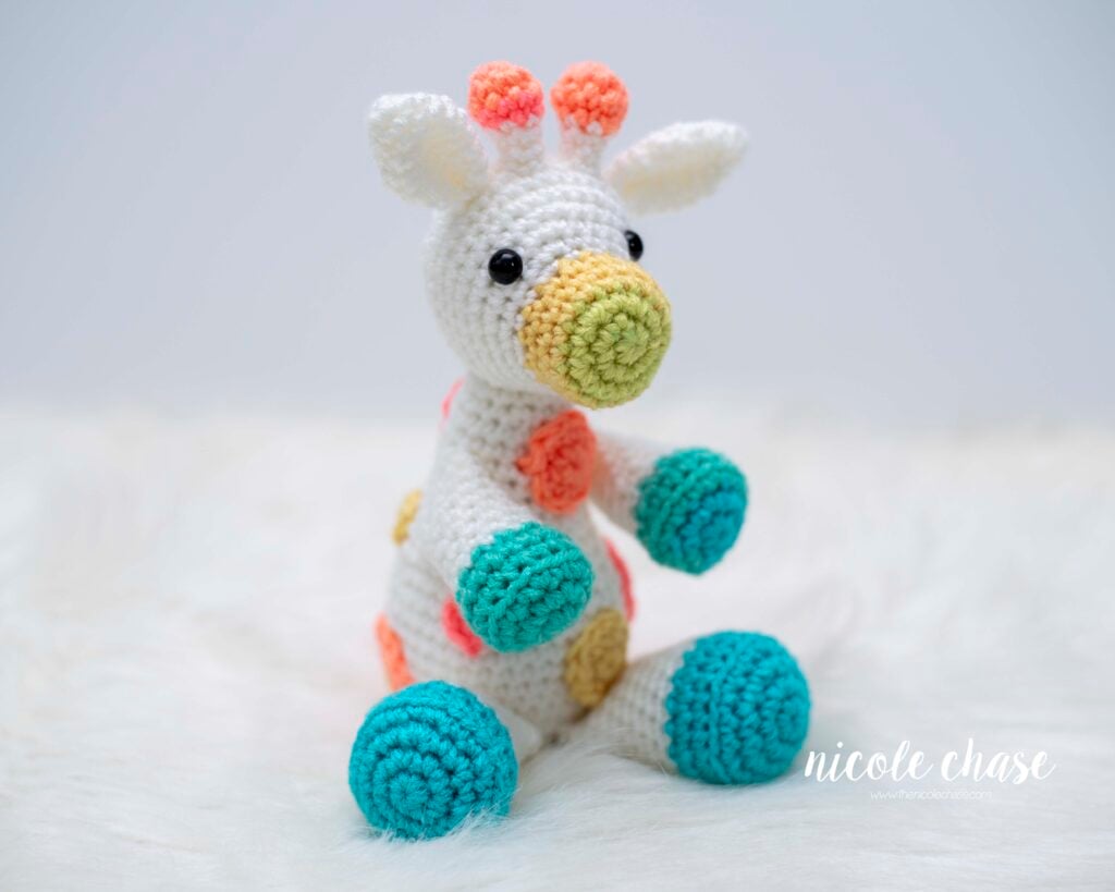 A rainbow version of Gerald the Giraffe made with white yarn and colorful accents. This free giraffe crochet pattern variation features teal hooves, orange and yellow spots, and a multicolored snout and horns, shown in a seated position. 