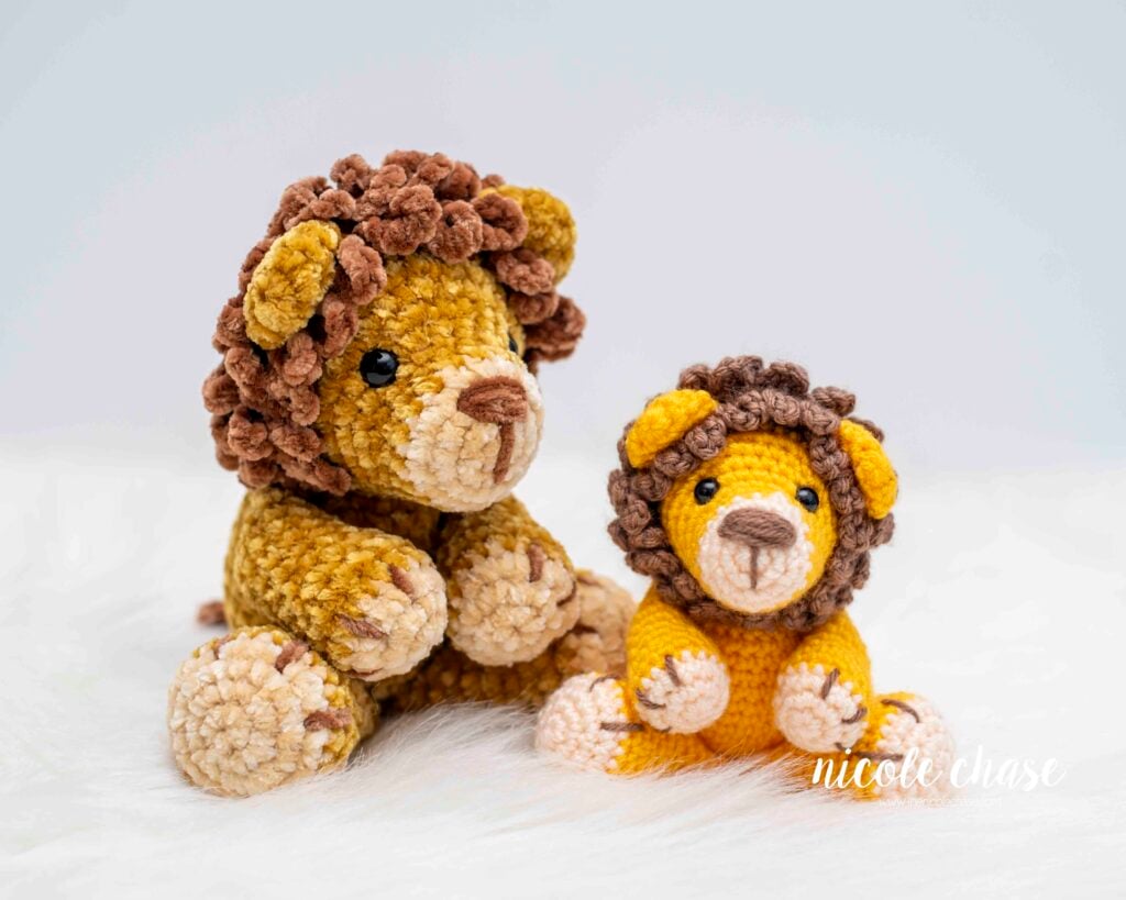 Two beginner crochet lions sitting on a white surface. The larger lion is made from velvet yarn with a dark brown loopy mane, while the smaller one is made from acrylic yarn with a similar brown mane. Both feature cream colored muzzles and black safety eyes. 