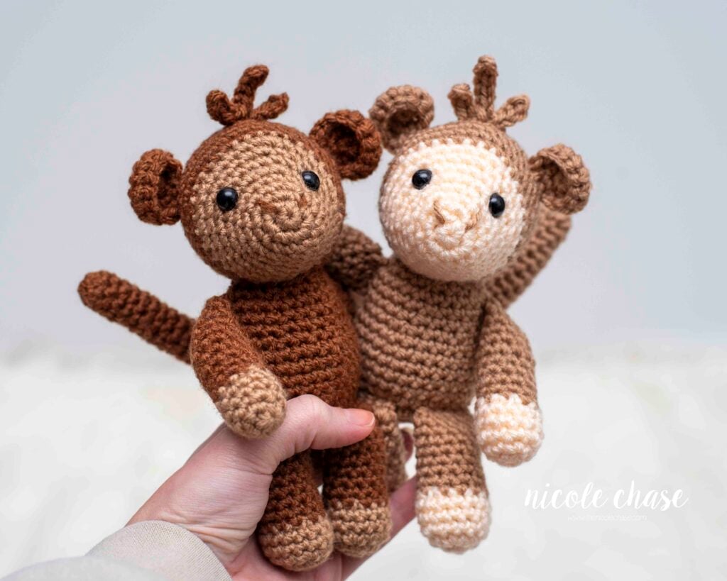 Two crochet monkeys being held. One is dark brown with a tan muzzle and paws, and the other is light brown with a cream muzzle and paws. Both have long tails, small ears, a tiny tuft of hair on top, and friendly embroidered faces. 